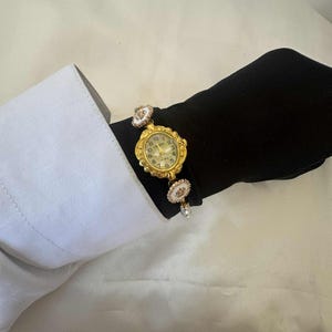 May include: A gold-toned wristwatch with an ornate, oval-shaped face and a cream-colored dial. The watch is attached to a bracelet with decorative white and gold accents. The watch is worn on a black sleeve, with a white shirt cuff visible.