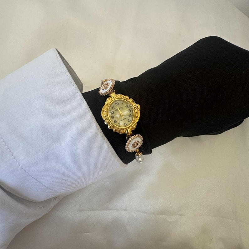 May include: A gold-toned wristwatch with an ornate, oval-shaped face and a cream-colored dial. The watch is attached to a bracelet with decorative white and gold accents. The watch is worn on a black sleeve, with a white shirt cuff visible.