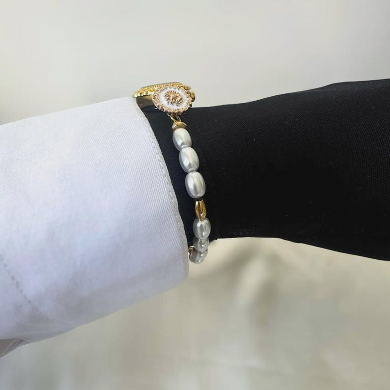 May include: A gold bracelet with a white and gold decorative clasp and a strand of white oval pearls. The bracelet is shown on a person's wrist, with a white shirt sleeve and a black sleeve in the background.