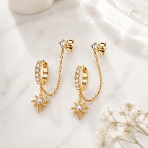 Peut inclure: Une paire de créoles dorées ornées d'une rangée de pierres transparentes. Chaque créole est reliée par une fine chaîne dorée à un clou d'oreille, avec une breloque en forme d'étoile à huit branches sertie d'une pierre centrale.