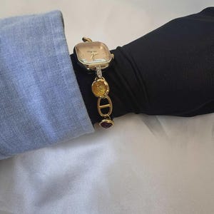 May include: A gold-toned wristwatch with a square face and a decorative band. The watch face is a light champagne color with gold hands and small crystals. The band features gold links, yellow circular accents, and a red gemstone.