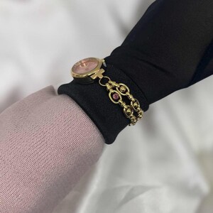 May include: A gold-toned wristwatch with a pink face and a matching gold bracelet. The bracelet features a double-strand design with small, round accents and pink gemstones. The watch is displayed on a black fabric.