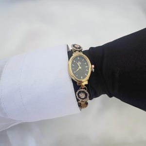 May include: Gold-toned oval watch with a black face and delicate gold hands. The watch features a gold band with decorative black and crystal accents. The watch is worn on a wrist with a white shirt cuff and a black sleeve.