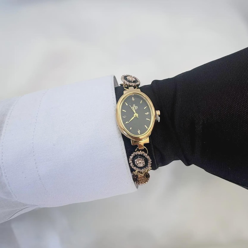 May include: Gold-toned oval watch with a black face and delicate gold hands. The watch features a gold band with decorative black and crystal accents. The watch is worn on a wrist with a white shirt cuff and a black sleeve.