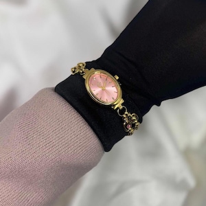 May include: A gold-toned wristwatch with a pink oval face and matching bracelet. The watch features delicate detailing and small pink gemstones. The watch is worn on a black sleeve, with a pink sweater visible in the background.