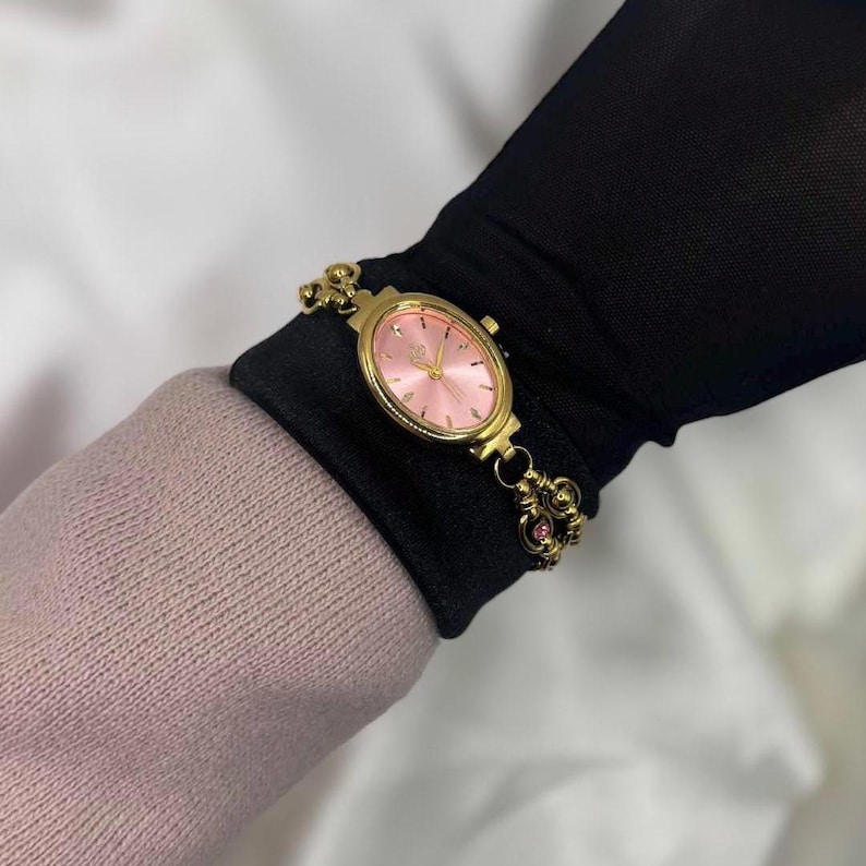 May include: A gold-toned wristwatch with a pink oval face and matching bracelet. The watch features delicate detailing and small pink gemstones. The watch is worn on a black sleeve, with a pink sweater visible in the background.