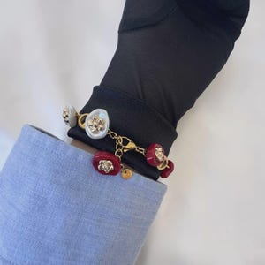May include: A gold-toned bracelet featuring alternating white and red decorative elements. The white elements have a floral design, while the red ones have a similar design. The bracelet is shown on a person's wrist, with a blue shirt sleeve in the background.