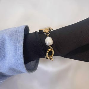 May include: A gold-toned watch with a white face and a pearl and chain bracelet. The watch is on a black sleeve, with a light blue sleeve in the background. The bracelet features a large, irregular white pearl.