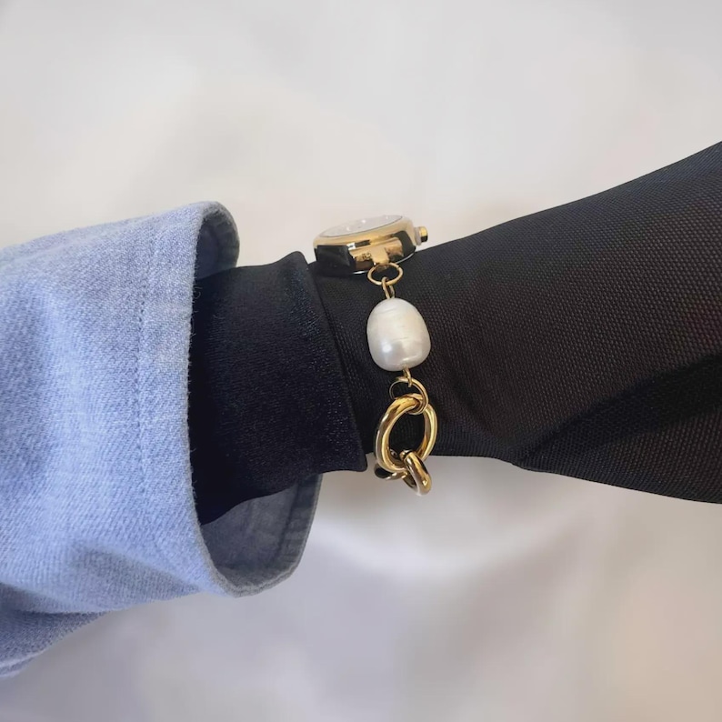 May include: A gold-toned watch with a white face and a pearl and chain bracelet. The watch is on a black sleeve, with a light blue sleeve in the background. The bracelet features a large, irregular white pearl.