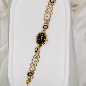 May include: Gold-toned wristwatch with a black oval face and a decorative floral bracelet. The bracelet features alternating black and white flower-shaped links with crystal accents. The watch is displayed on a white surface.