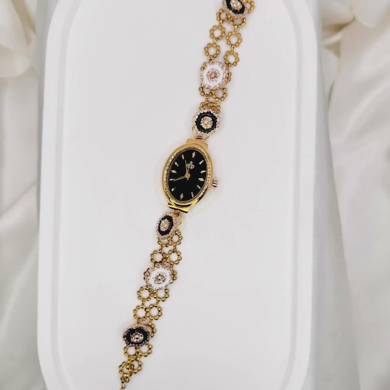 May include: Gold-toned wristwatch with a black oval face and a decorative floral bracelet. The bracelet features alternating black and white flower-shaped links with crystal accents. The watch is displayed on a white surface.