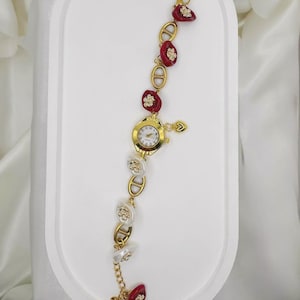 May include: Gold-toned wristwatch with a white clock face and gold hands. The watch band features alternating red and white decorative elements with gold accents. A small gold heart charm hangs from the watch.
