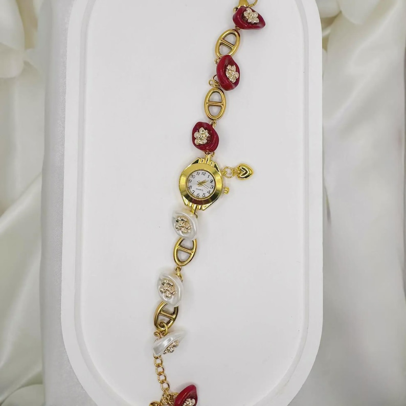 May include: Gold-toned wristwatch with a white clock face and gold hands. The watch band features alternating red and white decorative elements with gold accents. A small gold heart charm hangs from the watch.
