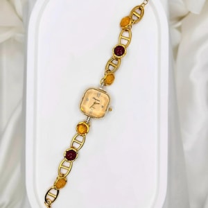 May include: A gold-toned wristwatch with a square face and a linked bracelet. The watch face is a light champagne color with black hands and Roman numerals. The bracelet features alternating gold links, amber-colored stones, and maroon-colored stones.