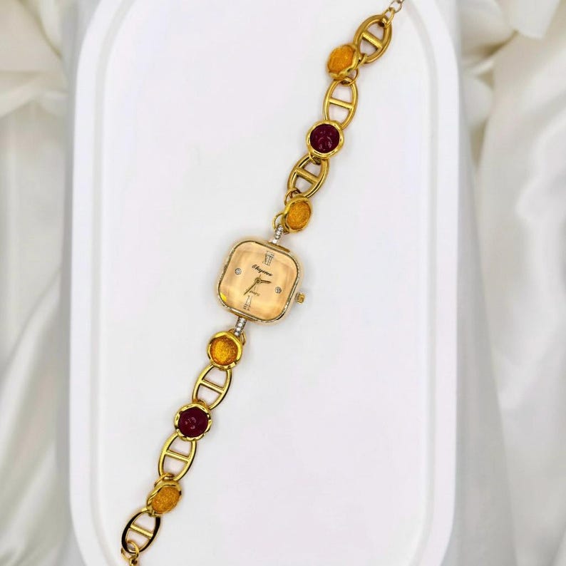 May include: A gold-toned wristwatch with a square face and a linked bracelet. The watch face is a light champagne color with black hands and Roman numerals. The bracelet features alternating gold links, amber-colored stones, and maroon-colored stones.