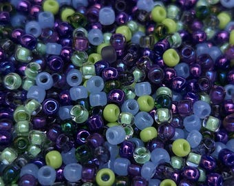 Pode incluir: Um close-up de uma variedade de contas de sementes em tons de roxo, azul e verde. As contas são pequenas e redondas, e estão dispostas em um padrão aleatório.