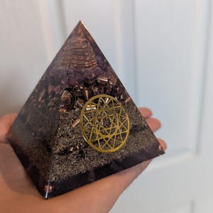 May include: A handcrafted pyramid-shaped orgone device. The pyramid is made of a clear resin with a dark purple and brown interior, featuring metal shavings and a gold-coloured geometric design. A copper coil sits at the top.