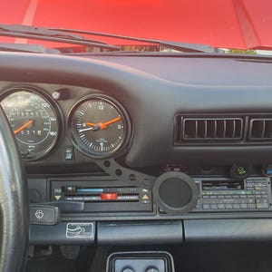 Könnte beinhalten: Armaturenbrett eines Oldtimers mit schwarzen Anzeigen, einem Tachometer und einer Uhr. Das Armaturenbrett ist schwarz mit roter Oberseite. Das Lenkrad ist links sichtbar.
