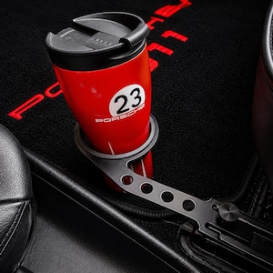 Puede incluir: Un termo rojo con tapa negra y el número 23, sujeto en un portavasos metálico. El termo tiene la palabra "PORSCHE" impresa. El portavasos está montado en el interior de un coche con asientos de cuero negro y alfombra negra.