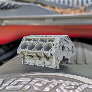 Peut inclure: Un modèle de bloc moteur gris imprimé en 3D. Le modèle comporte de multiples ouvertures cylindriques et des détails extérieurs. Le bloc moteur est présenté sur une surface sombre, avec un fond rouge visible.