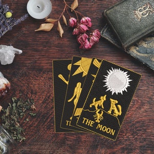May include: Four tarot cards on a wooden table with candles, crystals, herbs, and an old book. The cards are The Tower, The Star, The Sun, and The Moon. The cards are black with gold embroidery.