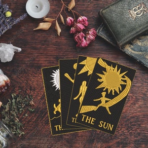 May include: Four tarot cards on a wooden table with a variety of objects, including candles, crystals, dried flowers, and herbs. The cards are black with gold embroidery and feature the symbols for the Sun, the Moon, the Star, and the World. The card in the foreground reads "THE SUN".