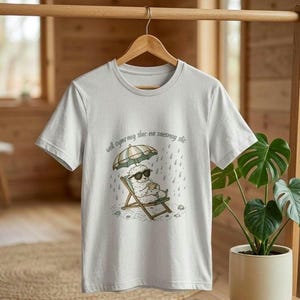 May include: A light gray t-shirt featuring a cartoon sheep wearing sunglasses and relaxing in a beach chair under an umbrella. The shirt has the text "well cymraeg slac na saesneg slic". The design includes rain and a cup.
