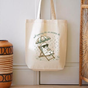 May include: Cream-colored tote bag featuring a cartoon sheep relaxing in a beach chair under an umbrella, wearing sunglasses and holding a cup. The bag has the text "well cymraeg slac na saesneg slic" printed on it.
