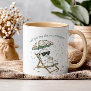 May include: White ceramic mug with a beige interior and handle. The mug features a cartoon sheep wearing sunglasses, relaxing in a beach chair under an umbrella in the rain. Text in a script font is above the sheep. The mug is on a burlap cloth.