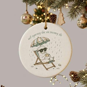 May include: Round Christmas ornament with a whimsical illustration of a sheep in sunglasses relaxing in a beach chair under an umbrella. The ornament has text in a script font and is hanging from a gold string.