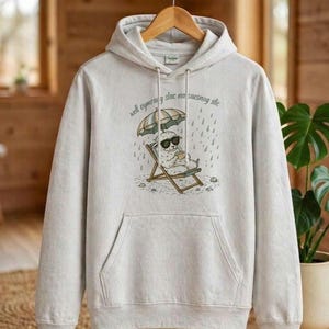 May include: Light gray hoodie with a kangaroo pocket and a drawstring hood. The front features a graphic of a sheep wearing sunglasses, sitting in a beach chair under an umbrella in the rain, with the text "well cymraeg slac na saesneg stic".
