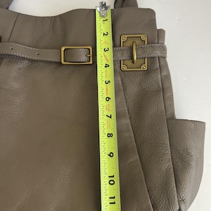 May include: Taupe leather handbag with a buckle closure and a side pocket. The bag is approximately 12 inches tall, measured with a yellow tape measure. The bag has a braided handle and gold-colored hardware.