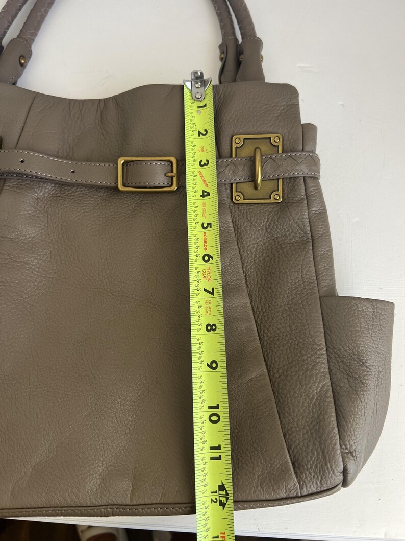 May include: Taupe leather handbag with a buckle closure and a side pocket. The bag is approximately 12 inches tall, measured with a yellow tape measure. The bag has a braided handle and gold-colored hardware.