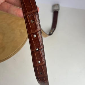 Puede incluir: Un cintur&oacute;n de cuero marr&oacute;n con un patr&oacute;n de cocodrilo. El cintur&oacute;n tiene una hebilla plateada y varios agujeros para ajustar. El cuero tiene un aspecto texturizado.