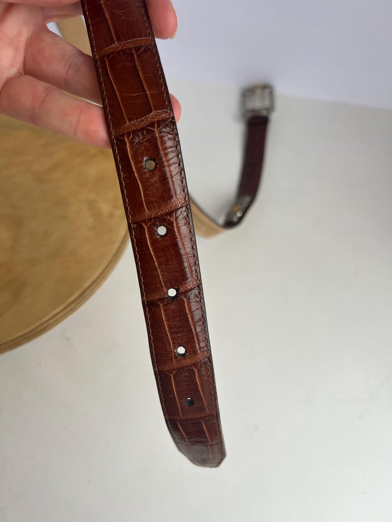 Puede incluir: Un cintur&oacute;n de cuero marr&oacute;n con un patr&oacute;n de cocodrilo. El cintur&oacute;n tiene una hebilla plateada y varios agujeros para ajustar. El cuero tiene un aspecto texturizado.