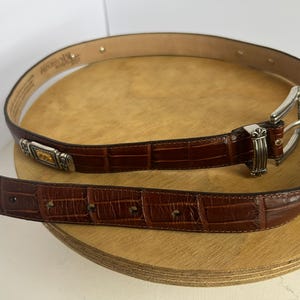 Puede incluir: Un cintur&oacute;n de cuero marr&oacute;n con textura de cocodrilo. El cintur&oacute;n tiene una hebilla plateada y detalles decorativos. El cintur&oacute;n mide aproximadamente 3,2 cm de ancho.