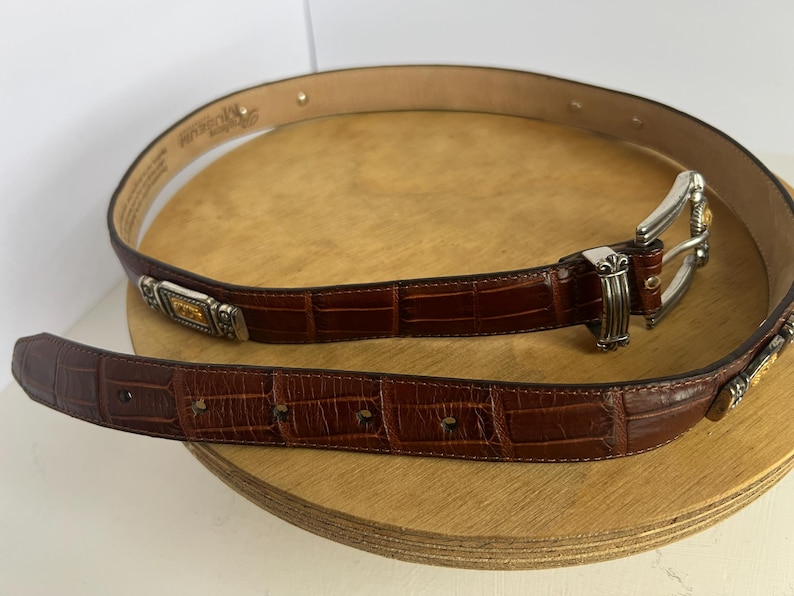 Puede incluir: Un cintur&oacute;n de cuero marr&oacute;n con textura de cocodrilo. El cintur&oacute;n tiene una hebilla plateada y detalles decorativos. El cintur&oacute;n mide aproximadamente 3,2 cm de ancho.