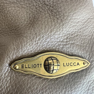May include: Close-up of a taupe leather bag with a textured surface. A gold-toned metal plaque is attached, featuring the brand name "ELLIOTT LUCCA" and a globe emblem.