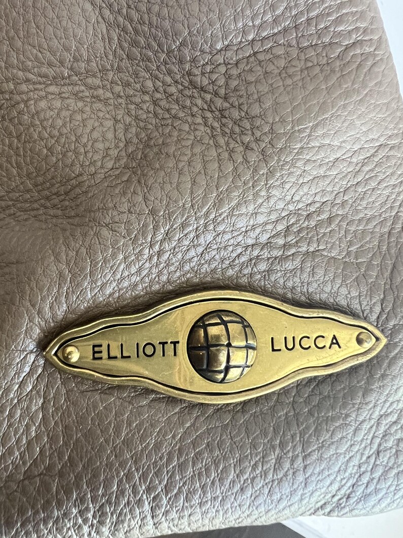 May include: Close-up of a taupe leather bag with a textured surface. A gold-toned metal plaque is attached, featuring the brand name "ELLIOTT LUCCA" and a globe emblem.