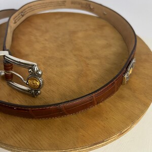 Puede incluir: Un cintur&oacute;n de cuero marr&oacute;n con textura de cocodrilo. El cintur&oacute;n tiene una hebilla plateada con detalles dorados y un detalle dorado decorativo cerca del lazo del cintur&oacute;n. El cintur&oacute;n se muestra sobre una superficie de madera.