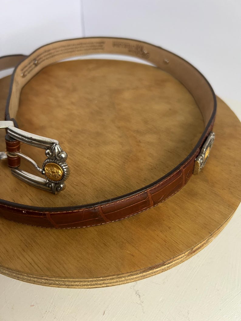 Puede incluir: Un cintur&oacute;n de cuero marr&oacute;n con textura de cocodrilo. El cintur&oacute;n tiene una hebilla plateada con detalles dorados y un detalle dorado decorativo cerca del lazo del cintur&oacute;n. El cintur&oacute;n se muestra sobre una superficie de madera.