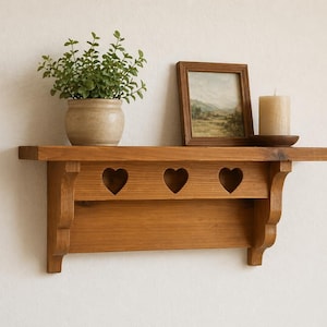 May include: A wooden shelf with a warm, honey-toned finish. The shelf features three heart-shaped cutouts and is adorned with a potted plant, a framed picture, and a candle on a wooden plate. The shelf is mounted on a white wall.
