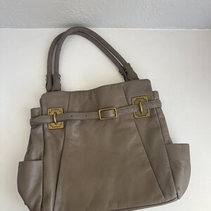 May include: Taupe leather handbag with braided handles, side pockets, and gold-tone hardware. The bag features a belt detail with a buckle closure and decorative metal clasps.