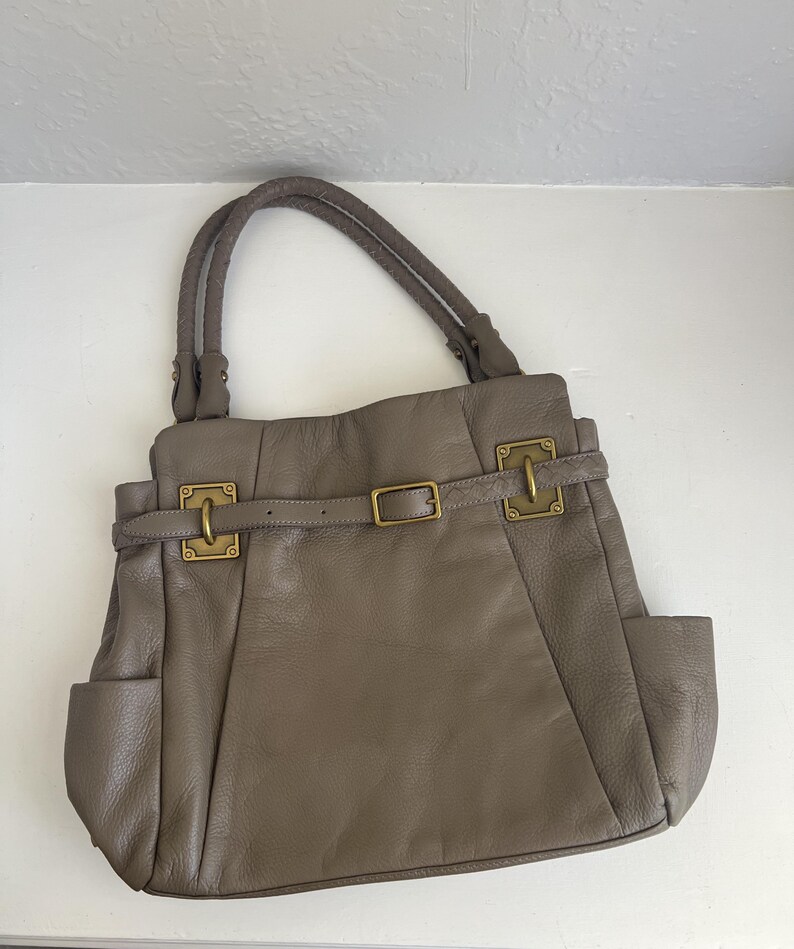 May include: Taupe leather handbag with braided handles, side pockets, and gold-tone hardware. The bag features a belt detail with a buckle closure and decorative metal clasps.