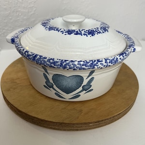 May include: A white ceramic casserole dish with a lid, featuring a blue heart design and blue floral accents. The dish sits on a round wooden trivet. The lid has a small knob for easy handling. The dish is designed for cooking and serving.