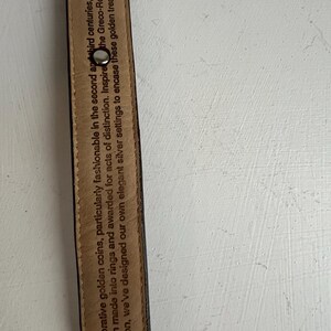 Puede incluir: Primer plano de un cintur&oacute;n de cuero negro con remaches plateados. El interior del cintur&oacute;n tiene texto impreso que dice: "Monedas de oro decorativas, particularmente de moda en los siglos II y III..."