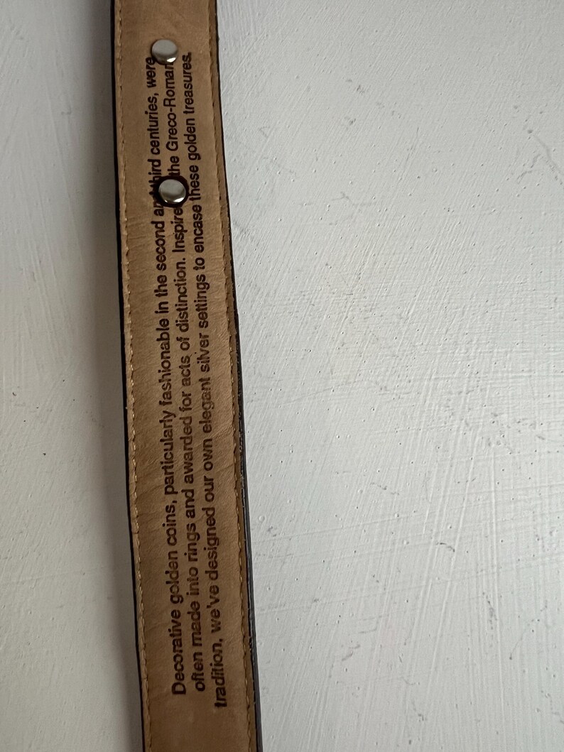 Puede incluir: Primer plano de un cintur&oacute;n de cuero negro con remaches plateados. El interior del cintur&oacute;n tiene texto impreso que dice: "Monedas de oro decorativas, particularmente de moda en los siglos II y III..."