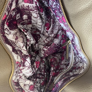 May include: Tan leather handbag with a gold zipper and hardware. The interior lining features a vibrant pattern of white, pink, and purple abstract shapes. The bag is open, revealing the patterned lining and a small zipper pocket.
