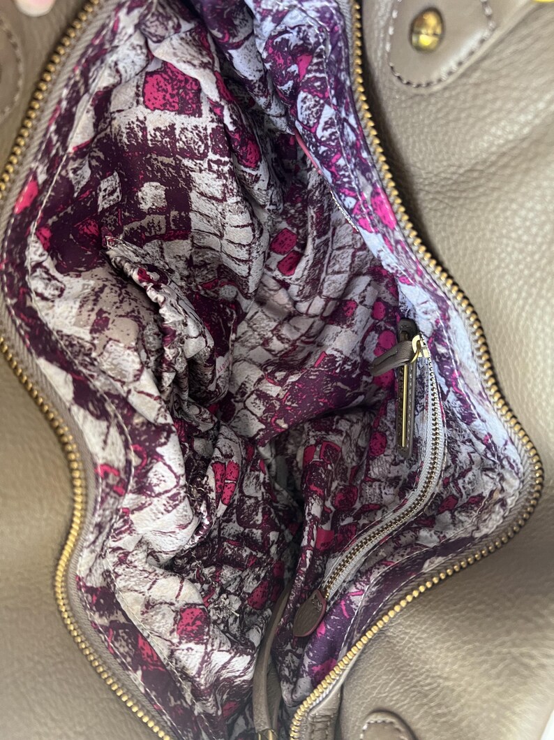 May include: Tan leather handbag with a gold zipper and hardware. The interior lining features a vibrant pattern of white, pink, and purple abstract shapes. The bag is open, revealing the patterned lining and a small zipper pocket.