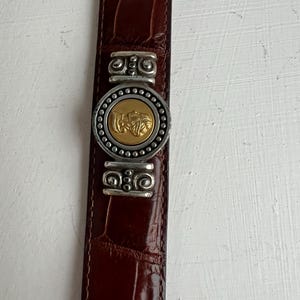 Puede incluir: Cintur&oacute;n de cuero marr&oacute;n con textura de cocodrilo. El cintur&oacute;n presenta una hebilla plateada con un medall&oacute;n dorado en el centro. La hebilla est&aacute; adornada con detalles decorativos plateados. Aproximadamente 3 cm de ancho.